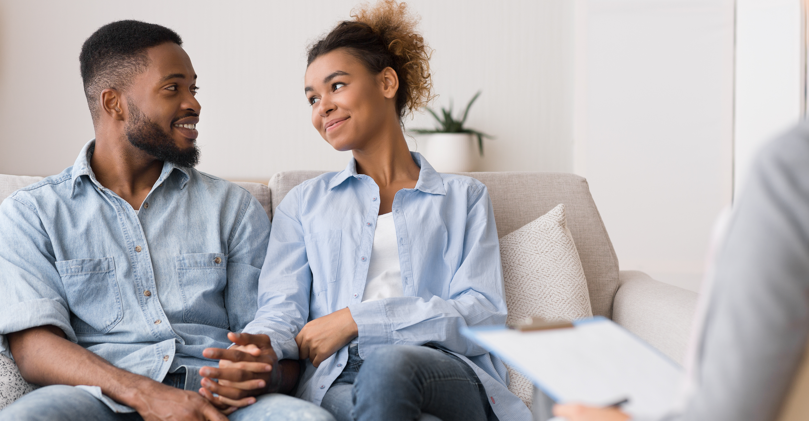 Young couple sitting on sofa, holding hands, black couple, casual dress, speaking with an advisor, relaxed, non-confrontational